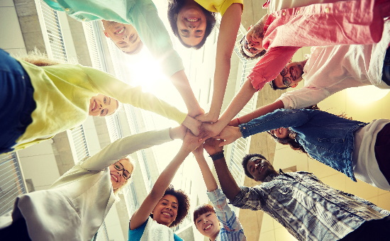 Die Kamera steht am Boden und schaut nach oben. Es sind Schüler:innen von unten zu sehen, unterschiedlichen Geschlechts, Kleiderfarbe und Hautfarbe, die im Kreis stehen und ihre Hände glücklich aufeinander legen.