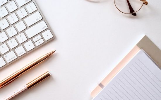 EIn Keyboard, Stifte, eine Brille und ein Notizblock in weiß und rosegoldenen Farben