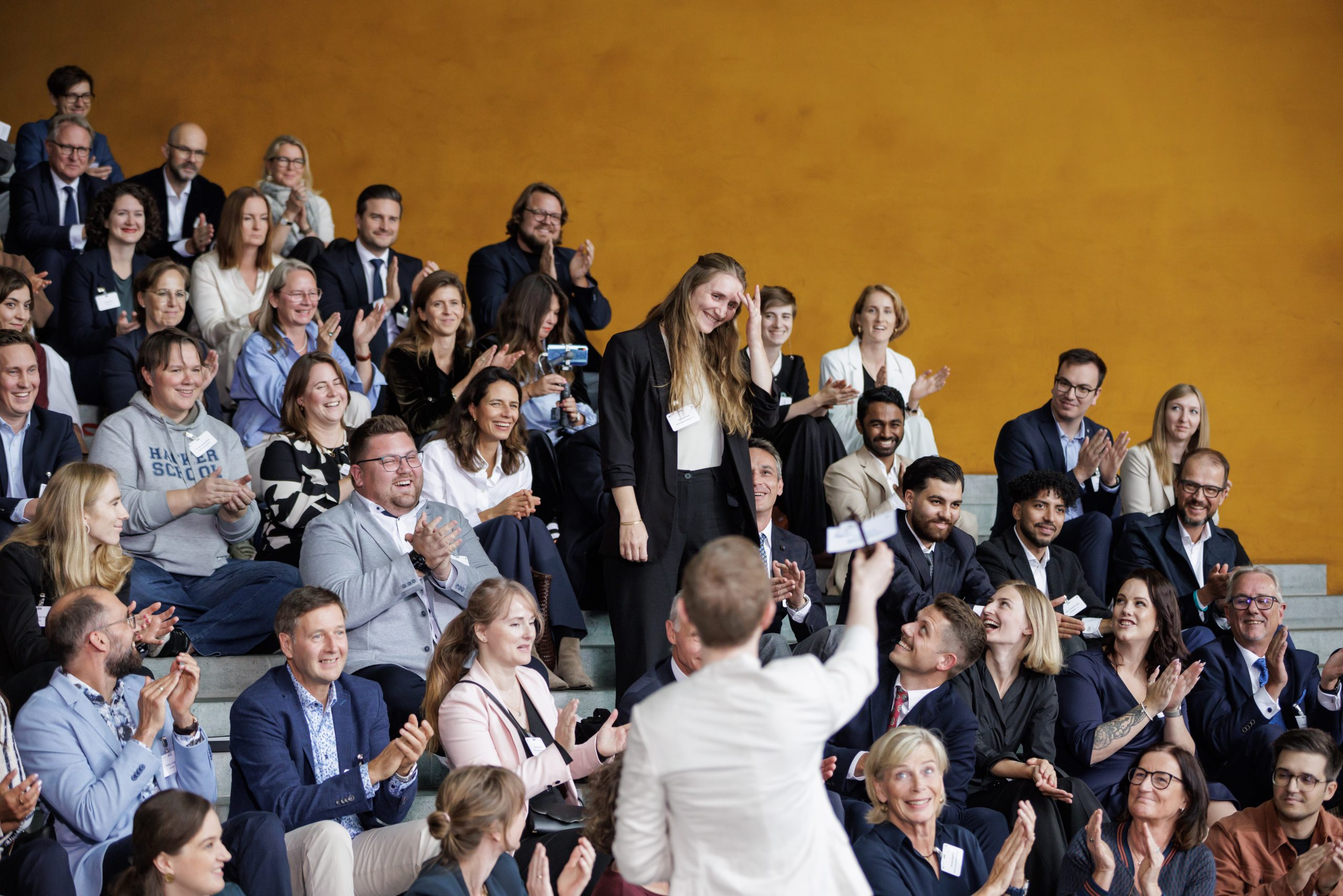 Die Projektmanagerin von Zeichen gegen Mobbing Vanessa steht in einer Menge von sitzenden Personen, die für sie klatschen.