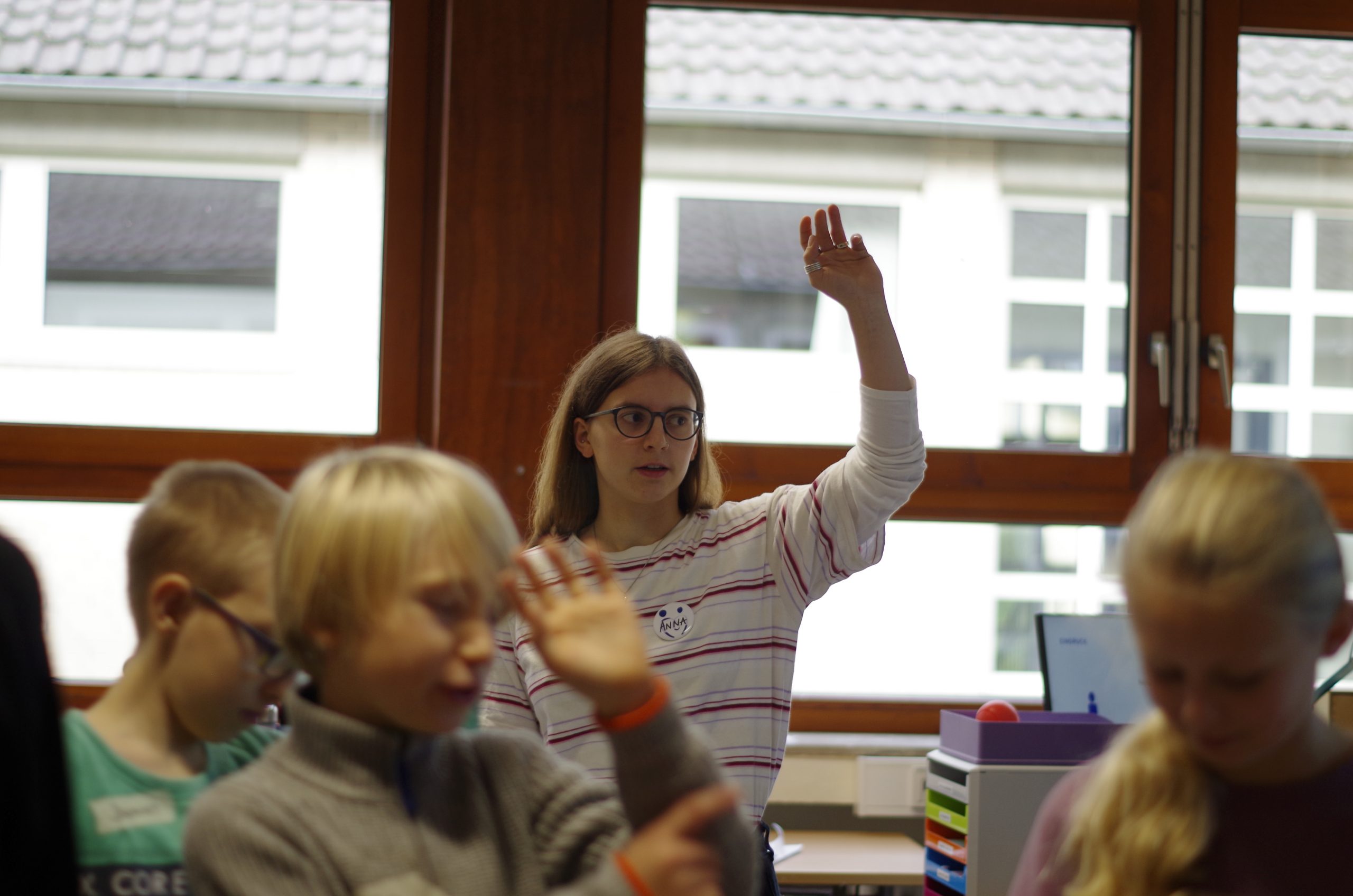 Eine junge Social Visionary leitet eine Schüler:innen-Gruppe an. Sie hebt die Hand während auch alle Schüler:innen die Hand heben.