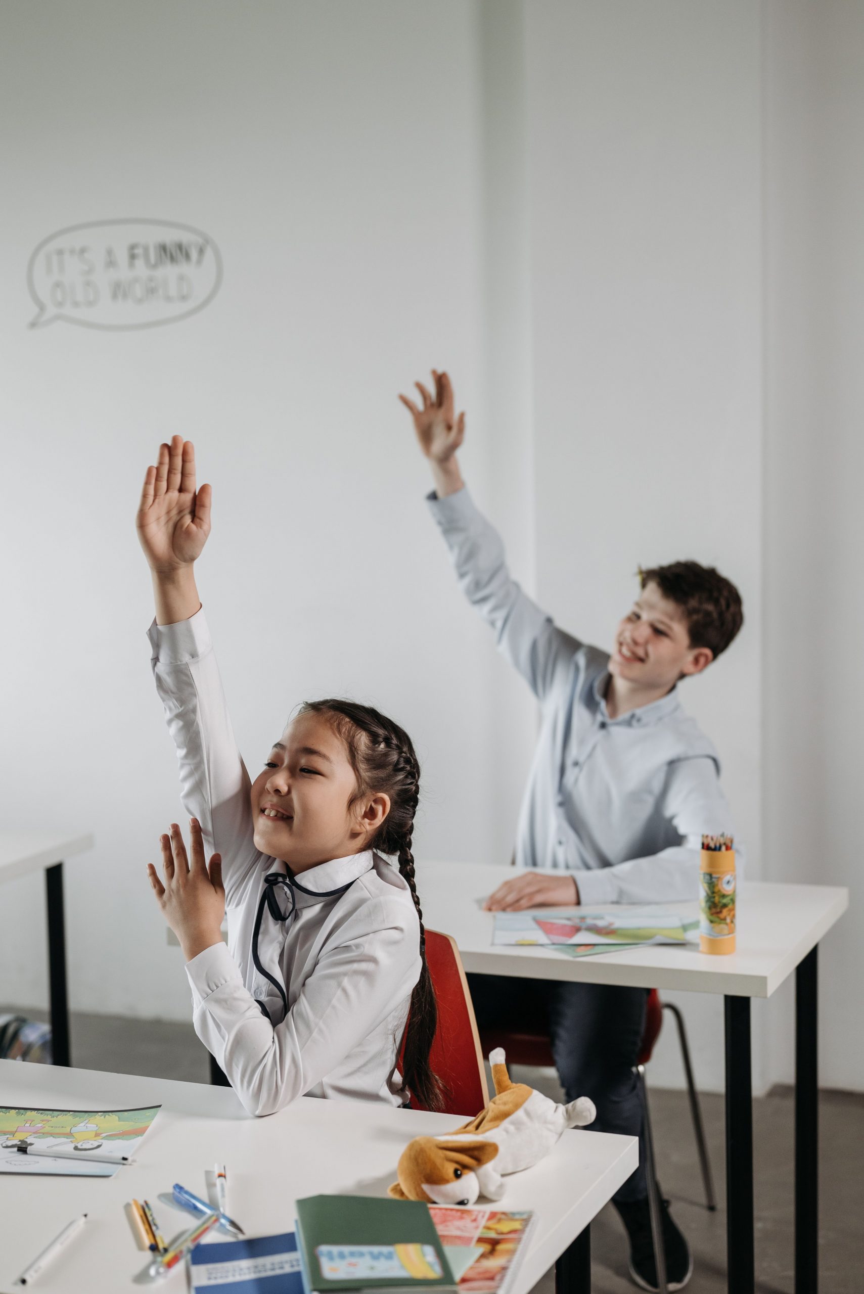 Zwei Kinder sitzen hintereinander an Tischen in der Schule. Beide melden sich, in dem sie den Arm heben. Beide lächeln.