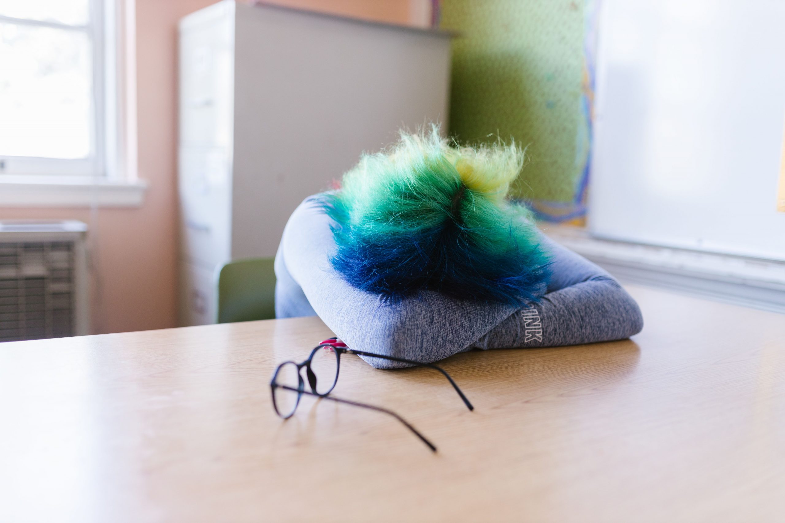 Eine Person mit bunten Haare hat den Kopf auf den Tisch gelegt, die Brille liegt daneben.