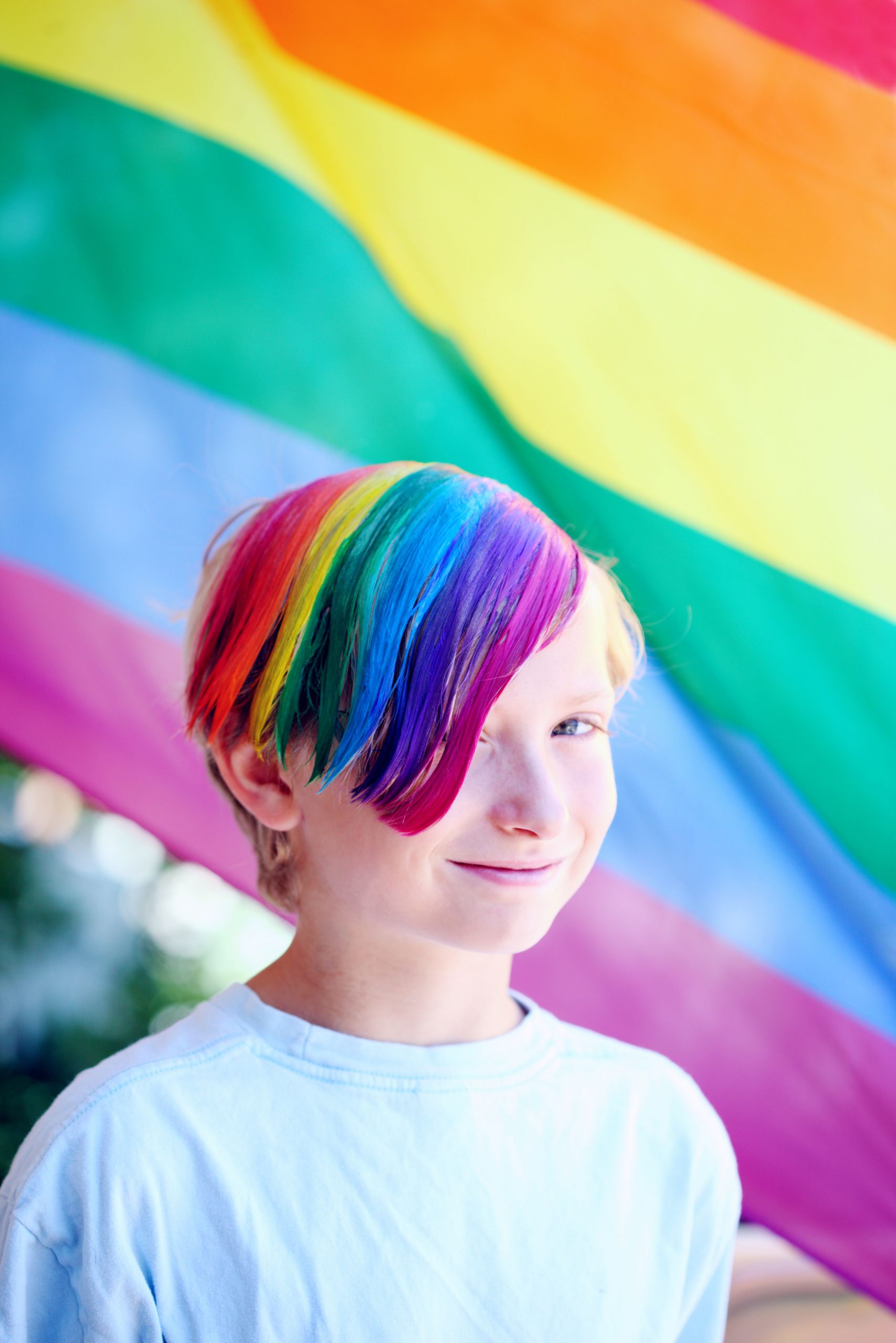 Ein Kind steht vor einer Regenbogenflagge und hat bunt gefärbte Haare in Farben des Regenbogens.